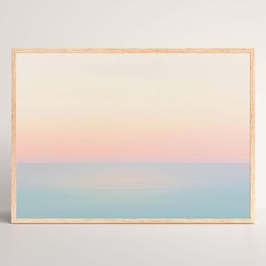 Puede incluir: Una impresión enmarcada que presenta un sereno paisaje marino. La obra de arte muestra un cielo degradado que transita del amarillo pálido al rosa suave, encontrándose con un tranquilo océano azul. El marco de madera añade un toque natural a la escena.