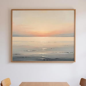 May include: A framed landscape painting depicts a serene ocean scene at sunset. The artwork features soft hues of peach, pink, and gray, with gentle waves and a warm, glowing sky. The painting is encased in a natural wood frame.