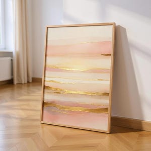 May include: Framed abstract art print. The artwork displays layers of soft pink, peach, and cream colours with gold foil accents. The piece is displayed on a wooden floor, leaning against a white wall.