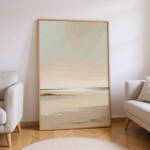 Può includere: Grande dipinto di paesaggio astratto incorniciato. L'opera presenta una scena serena con una tavolozza di colori beige chiaro e blu. Il dipinto è in una cornice di legno naturale e si appoggia a una parete bianca. L'opera è esposta vicino a un divano beige e un tappeto rotondo.