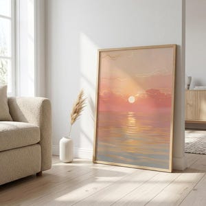 Peut inclure: Une impression d'art encadrée représente un coucher de soleil sur l'eau. L'œuvre présente une palette de couleurs chaudes de roses, d'oranges et de jaunes, avec le soleil se reflétant sur l'eau. Le cadre est en bois clair.