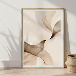 Może przedstawiać: Oprawiony abstrakcyjny wydruk artystyczny z płynącymi, organicznymi kształtami w odcieniach beżu i brązu. Grafika jest prezentowana na białej ścianie, z doniczką z wężownicą i wiklinowym koszem z kocem na pierwszym planie.