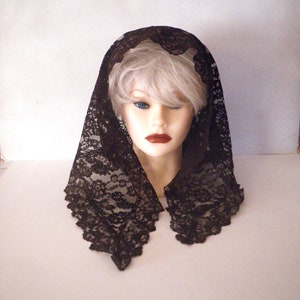 May include: A black lace veil draped over a mannequin head. The veil is intricately patterned with floral designs and has a scalloped edge. The mannequin has pale skin, blue eyes, and red lipstick.