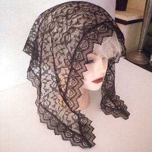 May include: A black lace head covering with a scalloped edge, displayed on a mannequin head. The lace features a floral pattern and a heart-shaped design along the border. The head covering is draped over the head and shoulders.