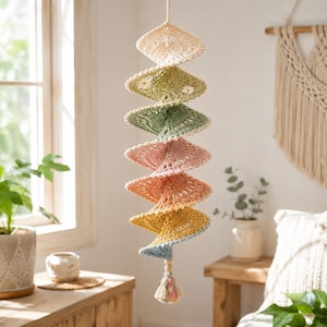 Puede incluir: Un móvil en espiral colgante hecho de hilo de ganchillo en un degradado de crema, verde salvia, rosa empolvado, melocotón, amarillo y azul claro. La estructura presenta intrincados patrones de encaje, pequeñas flores blancas y una borla.