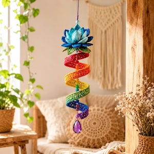 May include: A hanging crochet ornament featuring a blue lotus flower at the top, followed by a spiral tail in rainbow colours including red, orange, yellow, green, and blue. The spiral is accented with small colourful beads and a purple crystal drop.