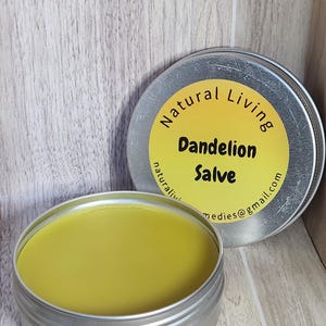 May include: Two open, silver-colored tins of dandelion salve. The salve is a light yellow color. The lid of one tin is visible, with a yellow label that reads "Natural Living Dandelion Salve". The email address "medies@gmail.com" is also printed on the label.