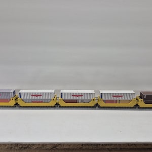Puede incluir: Un juego de trenes modelo con cinco vagones de mercancías. Los vagones amarillos transportan contenedores de envío en gris, blanco y marrón. Los contenedores tienen gráficos rojos y blancos. El tren está sobre una vía negra.