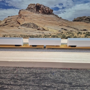 May include: Three white boxcars on a yellow flatbed train car, set against a backdrop of a desert landscape. The train cars are on a model railroad track, with a wooden surface in the foreground.