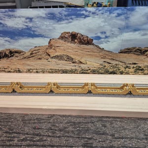 May include: A line of yellow model train flatcars with silver tracks. The flatcars are empty and have a detailed design. The background shows a desert landscape with a blue sky and clouds.