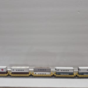Puede incluir: Un juego de trenes modelo con varios vagones de mercancías que transportan contenedores de envío. Los contenedores son blancos con varios logotipos y texto de la empresa, incluyendo "COSCO" y "CHINA SHIPPING". Los vagones son amarillos y están sobre una vía gris.