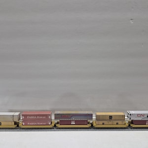 Puede incluir: Un tren modelo con varios vagones de carga que transportan contenedores de envío. Los contenedores son de varios colores, incluyendo rojo, amarillo y blanco, y presentan logotipos de empresas. El tren está sobre una vía, y el fondo es gris liso.