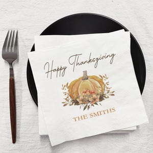 May include: White paper napkins with a watercolor pumpkin design and the text "Happy Thanksgiving" and "THE SMITHS" in brown script. The napkins are on a black plate with a silver fork.