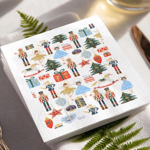 May include: A stack of white paper napkins with a festive Christmas design. The pattern includes nutcrackers, ballerinas, Christmas trees, wrapped gifts, and ornaments in red, blue, gold, and green.