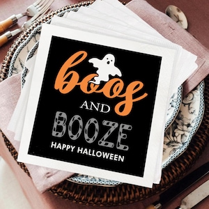 May include: Square white paper napkins with a black center featuring orange text that reads "Boos and Booze" with a white ghost graphic. The phrase "Happy Halloween" is printed below in white. Napkins are stacked on a decorative plate.