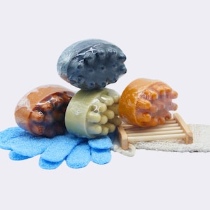 Puede incluir: Cuatro barras de jabón exfoliante en diferentes colores: marrón, verde, azul y naranja. Las barras están apiladas una encima de la otra y están sobre una toallita azul. Hay un jabonera de madera y una toallita blanca en el fondo.