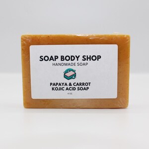 Puede incluir: Una barra rectangular de jabón artesanal, envuelta en plástico transparente. El jabón es de color naranja claro y tiene una etiqueta blanca con el texto "SOAP BODY SHOP HANDMADE SOAP" y "PAPAYA & CARROT KOJIC ACID SOAP". La etiqueta también indica 113 gramos.