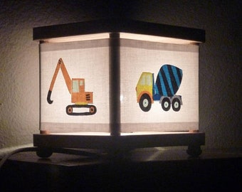 May include: A square table lamp with a wooden frame and a white fabric shade. The shade features illustrations of a yellow and orange excavator and a yellow and blue cement mixer truck. The lamp emits a soft glow from within the structure.