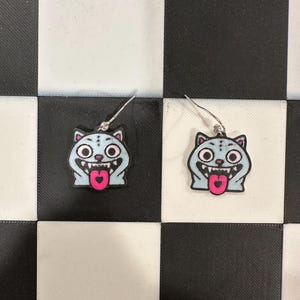 May include: A pair of earrings featuring a cartoon cat design. The earrings have a light blue cat face with a pink tongue, white teeth, and black and pink eyes. The earrings are on a black and white checkered surface.