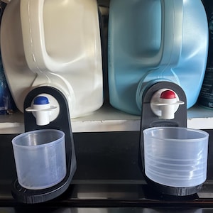 May include: Two large plastic jugs, one white and one light blue, with black dispensers and measuring cups attached. The white jug has a blue dispenser button, while the blue jug has a red one. The measuring cups are clear plastic.