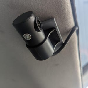 May include: A black car dash camera mounted to the ceiling of a vehicle. The camera has a small lens and a black plastic mount.