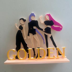 May include: A decorative shelf featuring three stylized figures in black outfits with colorful hair, standing above the word "GOLDEN" in yellow letters. The shelf is a light pink color and mounted on a white wall.