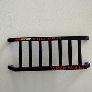 May include: Black ladder-style accessory with the text "TRD PRO" in red, yellow, and orange, and "Widdle Wadder" in red. The accessory has a ladder design.