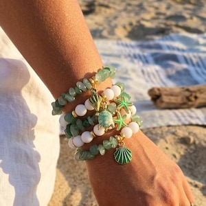 Op de afbeelding: Een verzameling armbanden met groene en witte kralen, versierd met goudkleurige bedels. De bedels zijn een zeeschelp, zeester en schildpad. De armbanden zijn op een pols gestapeld, met een strand en oceaan op de achtergrond.