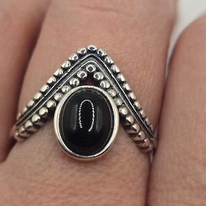 Peut inclure: Bague en argent ornée d'une pierre noire ovale sertie dans un anneau décoratif perlé. La bague présente un motif en forme de chevron au-dessus de la pierre, également perlé. La bague est portée sur un doigt.
