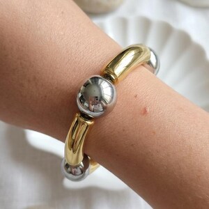 Könnte beinhalten: Ein elastisches Armband mit abwechselnd polierten, goldfarbenen, gebogenen Röhrenperlen und glatten, silberfarbenen Kugelperlen. Das Design besteht aus einem sich wiederholenden Muster aus metallischen zylindrischen und runden Elementen.