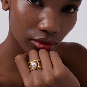 Könnte beinhalten: Ein goldfarbener Ring mit einer großen, oval geformten Perle. Der Ring hat ein strukturiertes, offenes Design. Der Ring wird an einem Finger getragen. Der Hintergrund ist neutral.