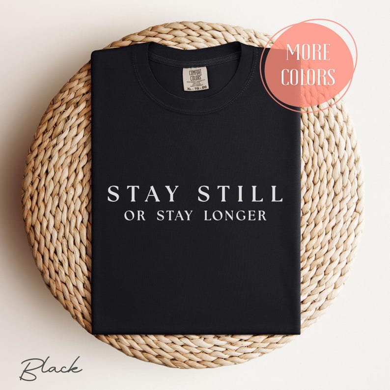 Folded black Comfort Colors t-shirt with white minimalist text reading “Stay Still or Stay Longer” — funny radiology quote shirt displayed on woven background.