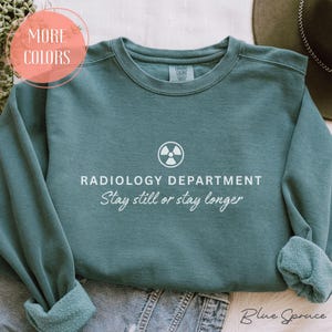 May include: Blue spruce-colored sweatshirt with the text "RADIOLOGY DEPARTMENT" and "Stay still or stay longer" in white. A white radiation symbol is above the text. The sweatshirt has a crew neck and long sleeves. The words "More Colors" are in a pink circle.