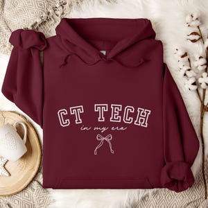 May include: A maroon hoodie with the text "CT TECH in my era" in white, along with a bow graphic. The hoodie has a drawstring hood and a front pocket. The garment is displayed on a soft, textured surface.
