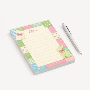 Peut inclure: Un bloc-notes avec un motif patchwork pastel, avec une page lignée crème avec le mot "Notes" en haut. Deux figurines de lapin avec des nœuds ornent le bloc-notes. Un stylo argenté repose à côté.