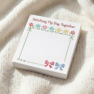 Peut inclure: Une pile de blocs-notes blancs avec un motif floral et nœuds au point de croix. Les blocs-notes sont décorés de fleurs et de nœuds colorés, et le texte "Stitching My Day Together" est imprimé en haut.