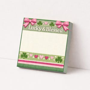 Może przedstawiać: Kwadratowy notatnik z zieloną obwódką i białym środkiem. Na górze różowe kokardy i napis "Lucky & Blessed". Projekt zawiera koniczyny i różowe serca. Dół ma różowo-biały pasiasty brzeg.