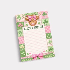 Puede incluir: Bloc de notas con un diseño de patchwork en rosa y verde, con tréboles y una vaca de las Tierras Altas con un lazo rosa. El bloc de notas tiene el texto "LUCKY NOTES" y un lazo rosa en la parte inferior.