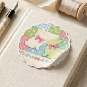 May include: An Easter card with two bunny rabbits wearing pink bows, set against a patchwork of pastel colours. The scalloped card sits on a linen-covered notebook, with a pen and a spool of ribbon in the background.