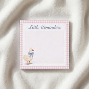 May include: A square notepad with a pink gingham border and the words "Little Reminders" in blue script. A watercolor illustration of a white goose with a blue bow is in the lower left corner. The notepad is on a white, textured surface.