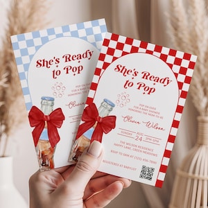 May include: Two baby shower invitations with the text "She's Ready to Pop." One invitation has a blue and white checkered background, the other has a red and white checkered background. Both feature a watercolor illustration of a bottle with a red bow.