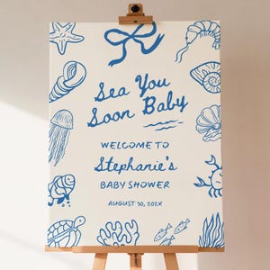 Puede incluir: Un letrero de baby shower color crema con ilustraciones náuticas azules de criaturas marinas y el texto "Sea You Soon Baby". El letrero da la bienvenida a los invitados al baby shower de Stephanie el 30 de agosto de 202X.