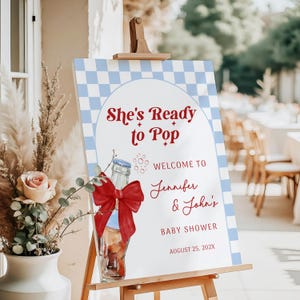以下が含まれることがあります： 木製のイーゼルに立てられたベビーシャワーのウェルカムサイン。青と白のチェック柄のボーダーがあり、赤いスクリプトで「She's Ready to Pop」というテキストが書かれています。赤いリボンが付いたボトルがサインに描かれています。ジェニファーとジョンのベビーシャワーにゲストを歓迎するサインです。