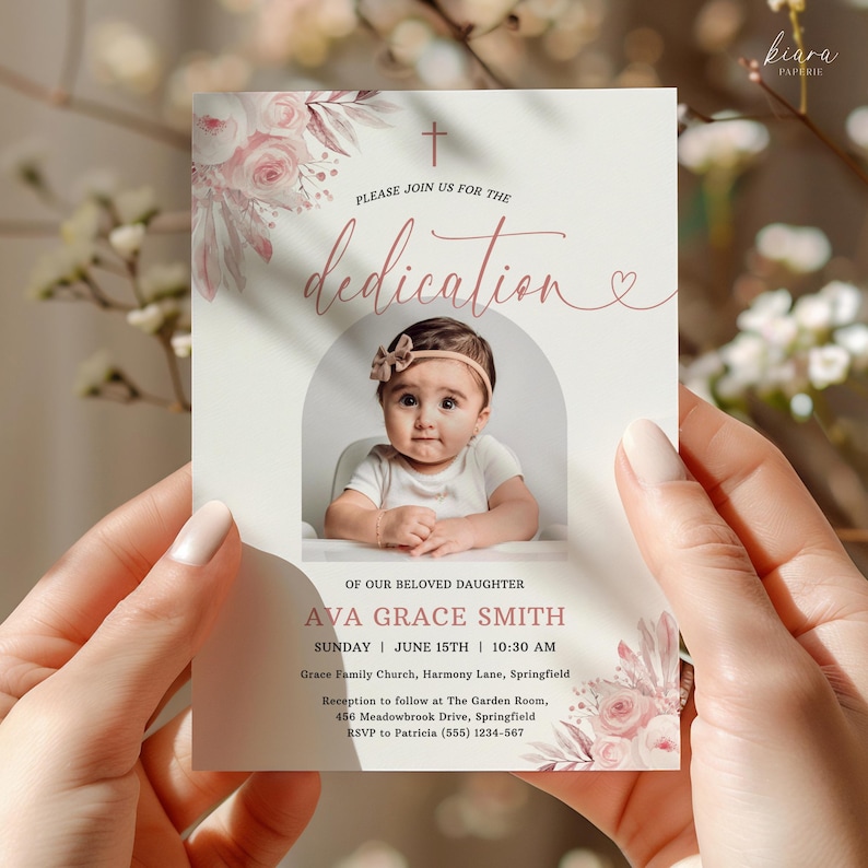 May include: A dedication announcement card held by a person. The card features a photo of a baby girl, floral accents, and the text "PLEASE JOIN US FOR THE dedication." Additional text includes the baby's name, date, time, and location.