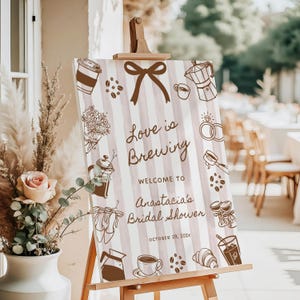 Puede incluir: Un letrero en un caballete de madera con el texto "Love is Brewing" y "Anastacia's Bridal Shower". El letrero tiene un fondo a rayas con ilustraciones de café en marrón. Hay un lazo en la parte superior. La fecha es el 29 de octubre de 202x.