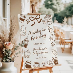 Könnte beinhalten: Ein weißes Schild auf einer Holzstaffelei mit braunen Illustrationen von Kaffeeartikeln und dem Text "A Baby is Brewing". Das Schild heißt die Gäste von Anastasias Babyparty willkommen, die für den 29. Oktober 202x geplant ist.