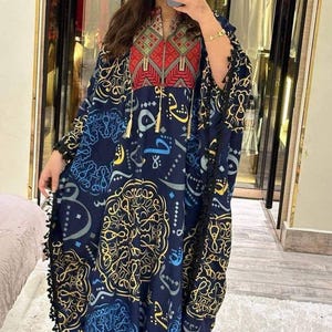 Op de afbeelding: Een marineblauwe kaftan met ingewikkelde gouden en blauwe patronen. De kaftan heeft een hoge kraag met rode en groene geometrische borduursels en kwastjes. De mouwen en zoom zijn afgezet met zwart kant.