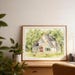 May include: A framed watercolour painting of a house with a light-coloured stone facade, a dark roof, and a red front door. The painting includes a green lawn, trees, and two small animals. The frame is made of light-coloured wood.