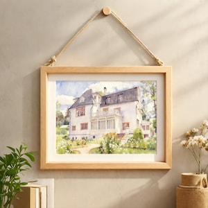 May include: A framed watercolour painting of a white house with a dark grey roof. The artwork shows a house with several windows, a porch, and a garden. The light wood frame is suspended by a natural rope, creating a decorative wall hanging.