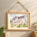 May include: A framed watercolour painting of a white house with a dark grey roof. The artwork shows a house with several windows, a porch, and a garden. The light wood frame is suspended by a natural rope, creating a decorative wall hanging.